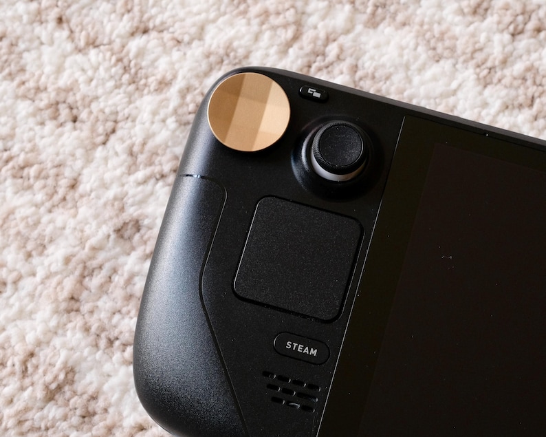 May include: Close-up of a black handheld gaming device with a gold-colored accent. The device features a joystick, a touchpad, and a button labeled "STEAM". The background is a textured, off-white surface.