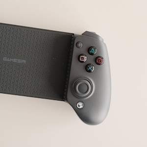 May include: A dark gray GAMESIR mobile gaming controller. The controller has a textured grip and a button layout with colored action buttons. The controller is designed for mobile gaming.