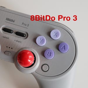 May include: A close-up of a grey 8BitDo Pro 3 controller. The controller has red joysticks, purple buttons labelled X, Y, A, and B, and a directional pad. The text "8BitDo Pro 3" is printed on the controller.