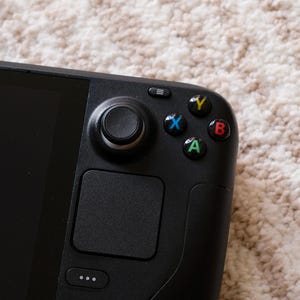 May include: Close-up of a black handheld gaming device. The device features a screen, a joystick, a touchpad, and colored buttons (green, blue, yellow, and red). The device is on a beige carpet.