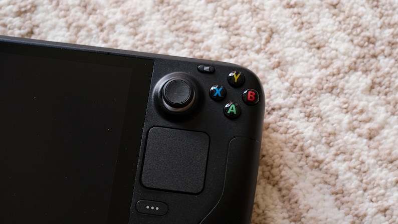 May include: Close-up of a black handheld gaming device. The device features a screen, a joystick, a touchpad, and colored buttons (green, blue, yellow, and red). The device is on a beige carpet.