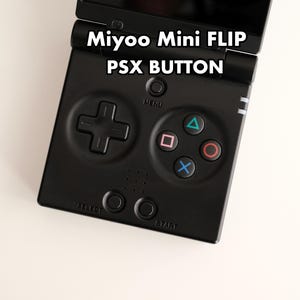 May include: A black Miyoo Mini FLIP handheld game console featuring the text Miyoo Mini FLIP PSX BUTTON. The device includes a directional pad, four action buttons with blue, pink, and red symbols, a menu button, and select and start buttons.