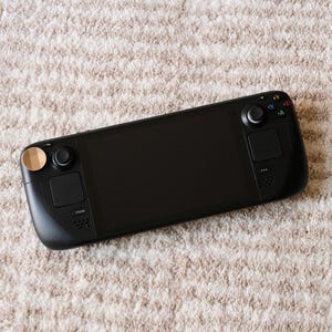 May include: A black handheld gaming console with a large screen, buttons, and joysticks. The console has a sleek design with a matte finish and is resting on a beige, textured surface.