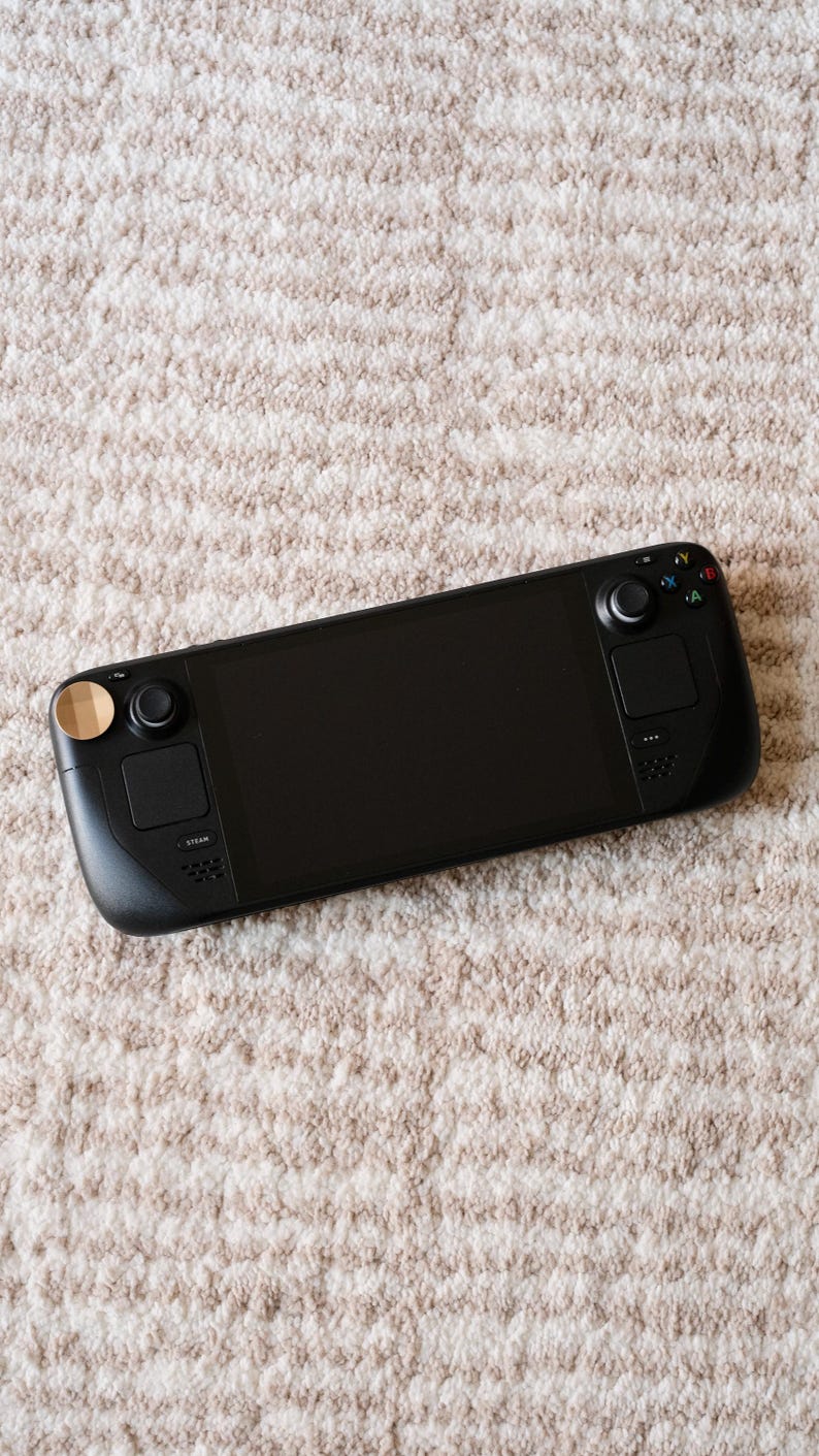 May include: A black handheld gaming console with a large screen, buttons, and joysticks. The console has a sleek design with a matte finish and is resting on a beige, textured surface.