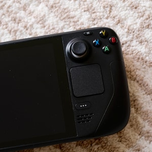 May include: A black handheld gaming console with a large screen and integrated controls. The console features a directional pad, analog stick, buttons, and a touchpad. The device is designed for portable gaming and entertainment.