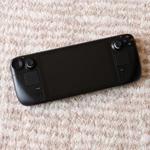 May include: A black handheld gaming console with a large screen, two control pads, and various buttons. The device is resting on a beige, textured surface, suggesting a soft rug or carpet.
