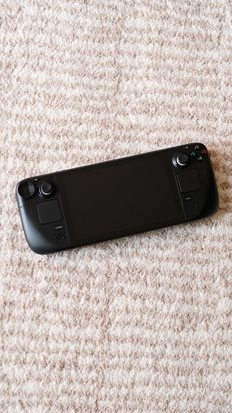 May include: A black handheld gaming console with a large screen, two control pads, and various buttons. The device is resting on a beige, textured surface, suggesting a soft rug or carpet.