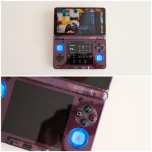 May include: A purple handheld gaming console with a dual-screen design. The top screen displays a game interface, while the bottom screen shows a user interface. The console features blue illuminated buttons and a directional pad. The console is open and ready to play.