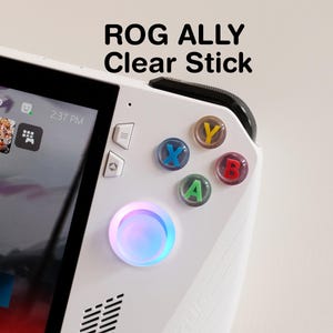 Può includere: Un dispositivo di gioco portatile ROG ALLY bianco con la scritta "ROG ALLY Clear Stick". Il dispositivo presenta pulsanti colorati, tra cui X, Y, A e B, e un pulsante circolare luminoso. Lo schermo mostra un'interfaccia di gioco.