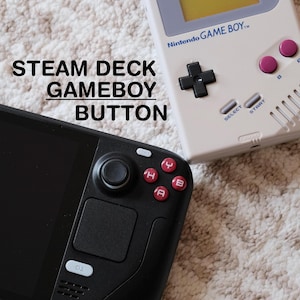 Könnte beinhalten: Ein schwarzer Steam Deck Controller mit roten Tasten mit den Buchstaben 'A', 'B', 'X' und 'Y' liegt neben einem weißen Nintendo Game Boy. Der Game Boy hat ein schwarzes Steuerkreuz, rosa Knöpfe und den Text 'Nintendo GAME BOY'. Der Text 'STEAM DECK GAMEBOY BUTTON' ist ebenfalls sichtbar.