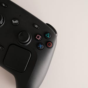 Może przedstawiać: Czarny bezprzewodowy kontroler do gier wideo z okrągłym padem kierunkowym, dużym analogowym drążkiem oraz czterema przyciskami akcji oznaczonymi niebieskim krzyżykiem, r&oacute;żowym kwadratem, zielonym tr&oacute;jkątem i czerwonym k&oacute;łkiem.