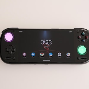 May include: A black handheld gaming device with a screen displaying the time and app icons. It has a pink and green illuminated button, a directional pad, and other control buttons. The device is on a white surface.