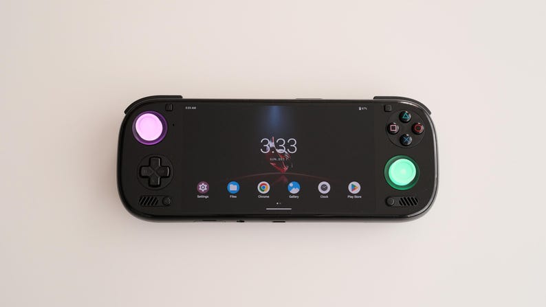 May include: A black handheld gaming device with a screen displaying the time and app icons. It has a pink and green illuminated button, a directional pad, and other control buttons. The device is on a white surface.