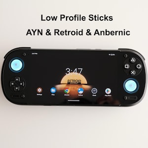 Puede incluir: Una consola de juegos portátil negra con joysticks de perfil bajo, una pantalla ancha que muestra la interfaz Retroid Pocket, un pad direccional y cuatro botones de acción. El texto Low Profile Sticks AYN & Retroid & Anbernic es visible.