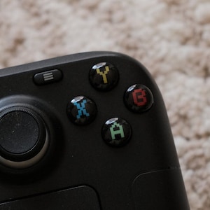 Könnte beinhalten: Ein schwarzer Gamecontroller mit vier Knöpfen, die mit A, B, X und Y beschriftet sind. Die Knöpfe sind schwarz mit farbigen Buchstaben und sind in einem Kreis um einen schwarzen Joystick angeordnet.