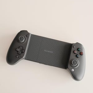 May include: A dark gray GAMESIR mobile gaming controller. The controller has a textured center section and two side grips with buttons and joysticks. The controller is designed for mobile gaming and has a sleek, modern design.