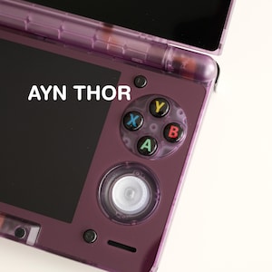 Könnte beinhalten: Eine lila Handheld-Spielkonsole mit dem Text "AYN THOR" auf dem Bildschirm. Die Konsole verfügt über ein Steuerkreuz, einen zentralen Steuerknüppel und farbige Aktionstasten. Das Gerät ist geöffnet und zeigt einen dunklen Bildschirm.
