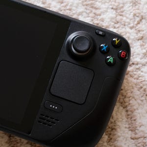 May include: Black handheld gaming console with a large screen and integrated controls. Features a joystick, directional pad, and colored action buttons. The device has a sleek, modern design.