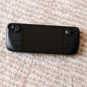 May include: A black handheld gaming console with a large screen, two control sticks, and various buttons. The device is lying on a beige, textured surface. The console has the word "STEAM" printed on it.