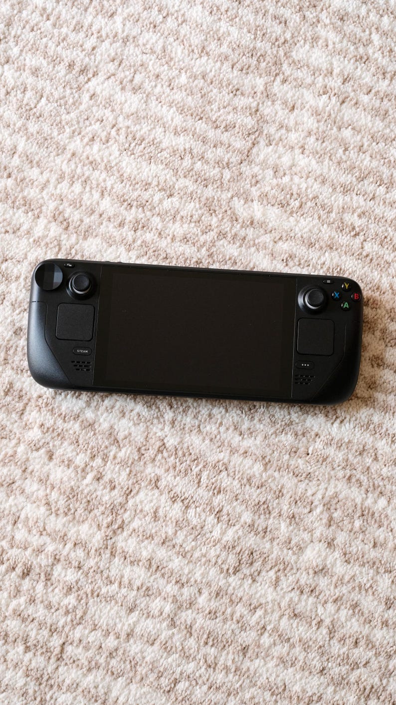 May include: A black handheld gaming console with a large screen, two control sticks, and various buttons. The device is lying on a beige, textured surface. The console has the word "STEAM" printed on it.