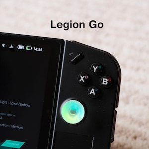 May include: A close-up of a black Legion Go handheld gaming device. The screen displays a menu with options. The right side features buttons labeled X, Y, B, and A, along with a glowing joystick. The text "Legion Go" is at the top.