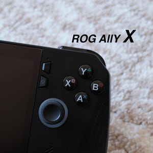 May include: Close-up of a black handheld gaming device with the text "ROG ALLY X" in black. The device features a screen, a directional pad, and colored action buttons: X, Y, A, and B. The background is a light-colored, textured surface.
