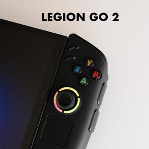 以下が含まれることがあります： 「LEGION GO 2」の文字が上部に表示された、黒色の携帯型ゲーム機のクローズアップ。画面と、カラフルなボタンとマルチカラーのジョイスティックを備えたコントローラーが特徴です。