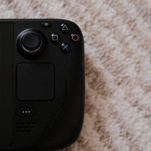 May include: A black handheld gaming console with a circular control pad, a square touchpad, and various colored buttons. The console is set against a neutral, textured background.