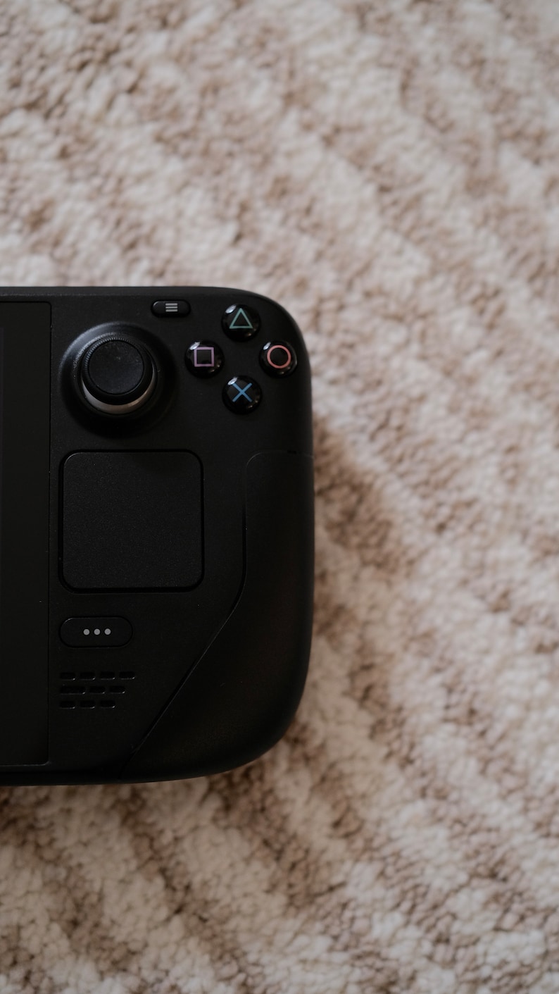 May include: A black handheld gaming console with a circular control pad, a square touchpad, and various colored buttons. The console is set against a neutral, textured background.