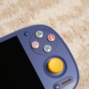 May include: Close-up of a handheld gaming device with a blue casing. It features a black screen and several colored buttons: green triangle, red circle, pink square, and blue cross. A yellow joystick is also visible.