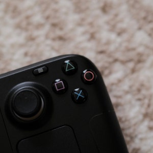 May include: Close-up of a black handheld gaming controller with a directional pad and action buttons. The action buttons are colored green, pink, blue, and purple. The controller has a black analog stick and a menu button.