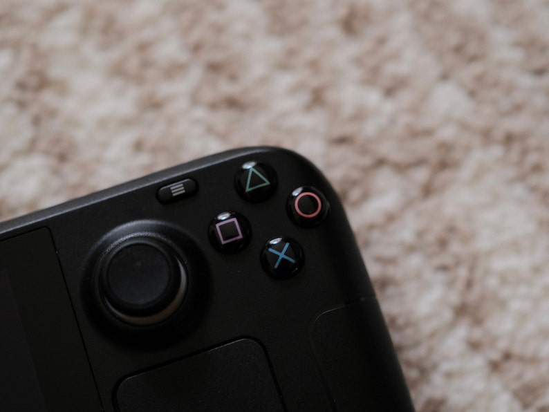 May include: Close-up of a black handheld gaming controller with a directional pad and action buttons. The action buttons are colored green, pink, blue, and purple. The controller has a black analog stick and a menu button.