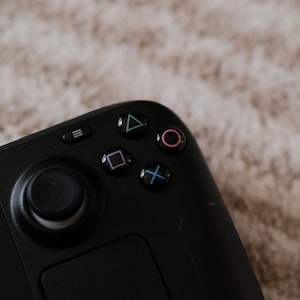 Könnte beinhalten: Ein schwarzer Gamecontroller mit einem runden Knopf, einem quadratischen Knopf, einem dreieckigen Knopf und einem runden Knopf. Der Controller hat ein Touchpad und einen Menüknopf.