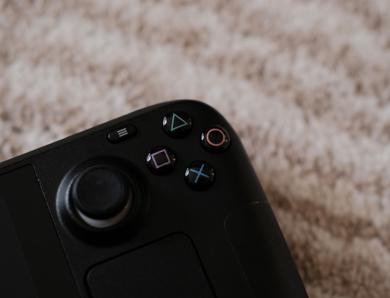 May include: Close-up of a black handheld gaming controller. The controller features a directional pad, a joystick, and action buttons with colored symbols: triangle, circle, square, and cross. The controller is set against a neutral background.