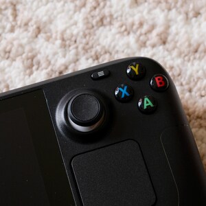 May include: Close-up of a black handheld gaming device. The device features a joystick, a touchpad, and colored buttons (green, blue, red, yellow). The device is on a textured, off-white surface.