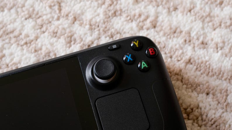 May include: Close-up of a black handheld gaming device. The device features a joystick, a touchpad, and colored buttons (green, blue, red, yellow). The device is on a textured, off-white surface.