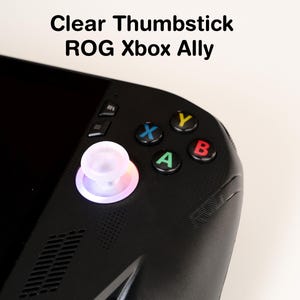 May include: Close-up of a black handheld gaming device with a clear thumbstick. The thumbstick is illuminated with a soft, colorful glow. The device has colored buttons: blue X, yellow Y, red B, and green A. Text on the image reads "Clear Thumbstick ROG Xbox Ally."