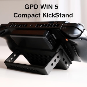 May include: Black GPD WIN 5 Compact KickStand. The stand is designed to hold a handheld gaming console. The stand has a kickstand design with a rectangular base and angled support legs. The text "GPD WIN 5 Compact KickStand" is printed on the top.