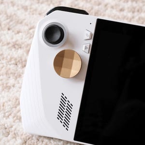 Puede incluir: Consola de juegos portátil blanca con pantalla negra y controles. La consola presenta un joystick negro, dos botones y un acento circular dorado. Se ven rejillas de ventilación anguladas en el lateral.