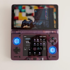 May include: A purple handheld gaming console with a screen displaying a video game interface. The console has buttons, joysticks, and a D-pad. The screen shows the text "60 FPS MODE" and "15 FPS".