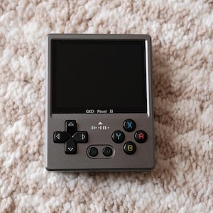 May include: A grey handheld gaming device with a black screen. The device has a directional pad, four face buttons, and a GKD Pixel II label.