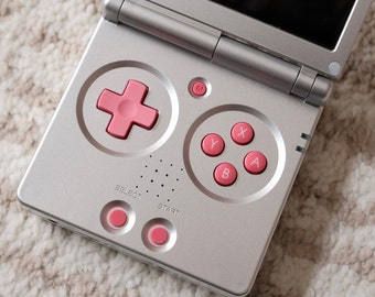 May include: Silver handheld gaming console with a pink directional pad and pink A, B, X, and Y buttons. The console has a small screen and a start and select button. The console is open and ready to play.