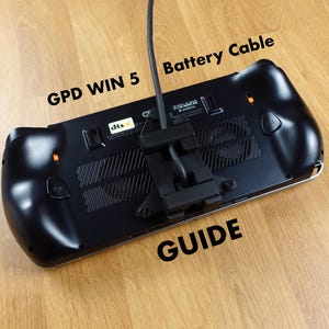 Op de afbeelding: Zwarte GPD WIN 5 handheld gameconsole met een batterijkabel bevestigd. De console ligt op een houten oppervlak, met de tekst "GPD WIN 5", "Battery Cable" en "GUIDE" zichtbaar.