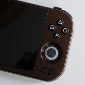 Può includere: Primo piano di un dispositivo di gioco portatile con un involucro marrone e uno schermo nero. Dispone di un pad direzionale, pulsanti di azione e un joystick. I pulsanti sono bianchi e neri. Progettato per il gioco portatile.