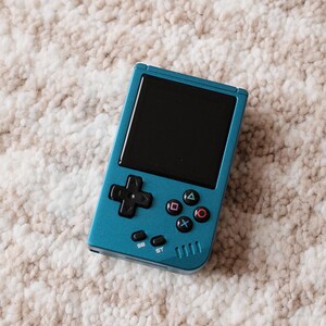 May include: A teal handheld gaming console with a black screen and black control buttons. The console has a directional pad, action buttons, and start/select buttons. The device is on a textured, off-white and tan surface.