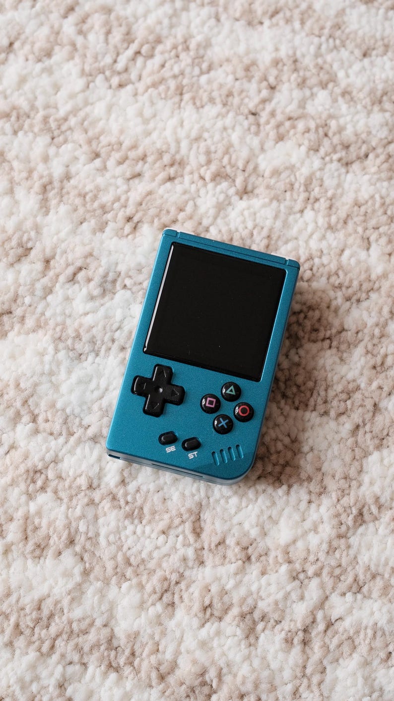 May include: A teal handheld gaming console with a black screen and black control buttons. The console has a directional pad, action buttons, and start/select buttons. The device is on a textured, off-white and tan surface.
