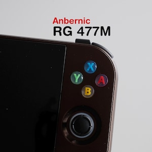 May include: Close-up of a brown handheld gaming console with the text "Anbernic RG 477M" in red and black. The console features colorful buttons: blue, red, green, and yellow. A black joystick is visible below the buttons.