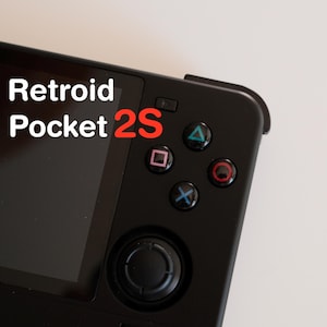 May include: Black handheld gaming device with a screen and control buttons. The device has a directional pad, action buttons, and a joystick. The text "Retroid Pocket 2S" is displayed on the screen.