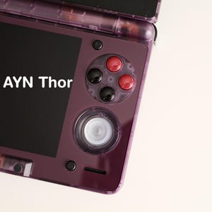 Puede incluir: Una consola de juegos portátil morada con el texto "AYN Thor" en la pantalla. El dispositivo tiene botones negros y rojos, un mando direccional y un control central. La pantalla es negra y el dispositivo está abierto.