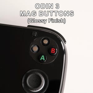 Pode incluir: Um dispositivo de jogo portátil preto com o texto ODIN 3 MAG BUTTONS (Glossy Finish). O dispositivo inclui um joystick circular preto e quatro botões de ação: dois botões metálicos prateados, um botão A verde e um botão B vermelho.